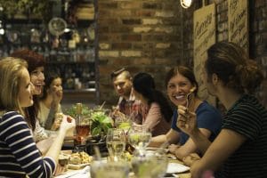 A group friends in there late 20s are socialising, eating and drinking together in a bar and restaurant. They are all dressed casual and smiling and laughing with one another.